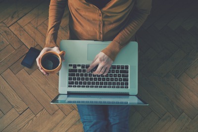 person sitting on floor with laptop in lap and coffee cup in one hand
