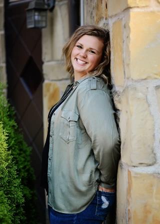 smiling young woman with hands in her back pockets leaning on stone wall