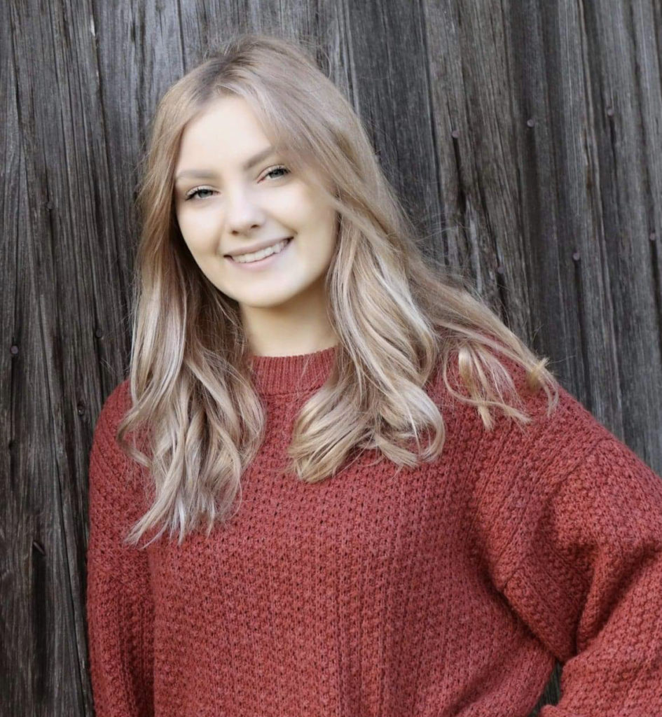 smiling young woman with red cable knit sweater in front of wooden wall