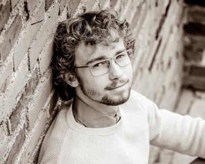 young man with glasses leaning up against a brick wall